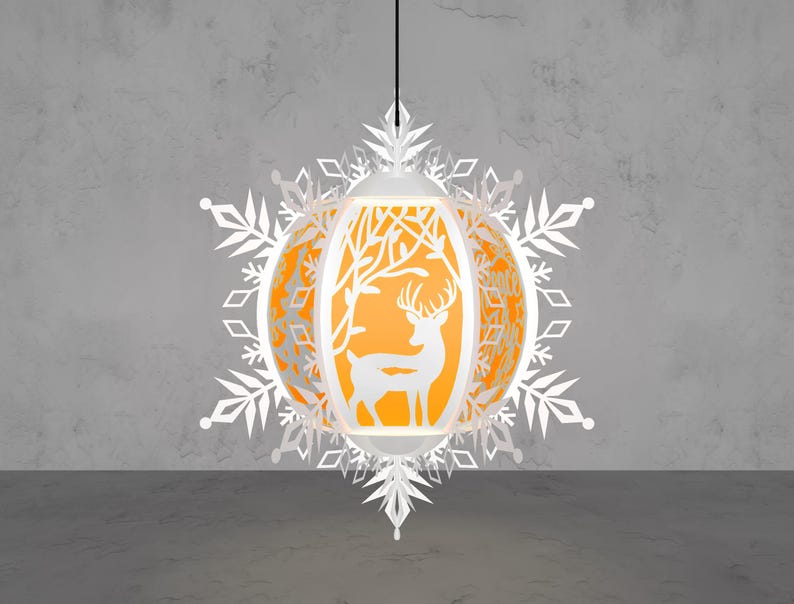 May include: A white paper lantern with a deer silhouette and tree design, surrounded by snowflake cutouts. The lantern has an orange interior and hangs from a black cord. The design is intricate and festive.