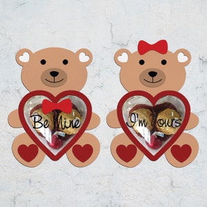 Peut inclure: Deux porte-friandises en forme d'ours en peluche brun pour la Saint-Valentin. Chaque ours tient un cœur rouge avec une fenêtre transparente. Le cœur du ours de gauche dit "Be Mine" et le cœur du ours de droite dit "I'm Yours".