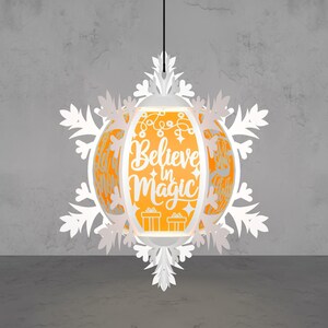 May include: A white paper lantern with a snowflake design, hanging from a black cord. The lantern has an orange interior with the words "Believe in Magic" and festive illustrations. The overall design is festive and decorative.