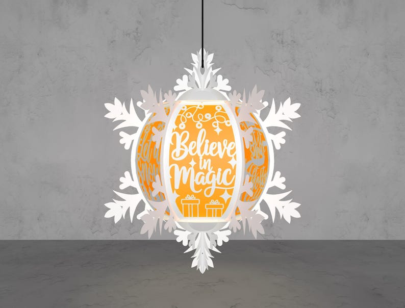 May include: A white paper lantern with a snowflake design, hanging from a black cord. The lantern has an orange interior with the words "Believe in Magic" and festive illustrations. The overall design is festive and decorative.
