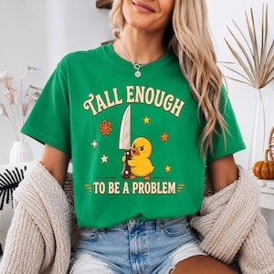 Könnte beinhalten: Ein leuchtend grünes Kurzarm-T-Shirt mit einer Grafik eines gelben Entchens, das ein großes Messer hält. Der Text TALL ENOUGH TO BE A PROBLEM steht in einer Retro-Schriftart, umgeben von kleinen orangefarbenen und gelben Stern- und Blumensymbolen.