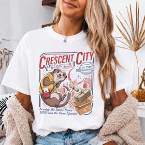 Può includere: Una t-shirt bianca con la grafica di una lontra che indossa un gilet giallo e porta una borsa postale rossa. Il testo recita Crescent City Postal Service, On Time Delivery e Serving the Istros River and the Bone Quarter con un pacco.