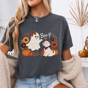 Könnte beinhalten: Ein anthrazitfarbenes T-Shirt mit einem Grafikdruck eines weißen Geistes mit karierter Schleife und eines weißen Hundes mit kariertem Halstuch. Das Design zeigt orangefarbene Kürbisse, Sonnenblumen und den Schriftzug Boo-P mit einem Herz.