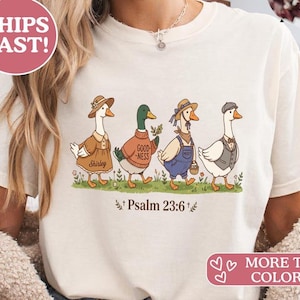 Könnte beinhalten: Cremefarbenes T-Shirt mit einer skurrilen Illustration von vier Gänsen in verschiedenen Outfits, die auf Gras laufen. Der Text "Psalm 23:6" steht unter den Gänsen. Das Shirt hat auch den Text "MORE TEE COLORS!"
