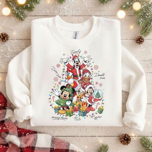 Könnte beinhalten: Weißes Sweatshirt mit einem farbenfrohen Weihnachtsmotiv von Disney-Figuren. Mickey, Minnie, Goofy, Donald, Daisy und Pluto sind in festlicher Kleidung dargestellt, umgeben von Lichtern und Geschenken. Die Namen der Charaktere sind darunter geschrieben.