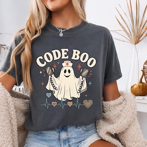 Op de afbeelding: Een antracietgrijs t-shirt met korte mouwen en een afbeelding van een geest met een verpleegsterskapje die defibrillatorpeddels vasthoudt. De tekst CODE BOO staat in crèmekleurige letters boven de geest, met een hartslaglijn en hartjes.