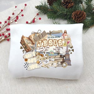 Könnte beinhalten: Weißes Sweatshirt mit einem Collage-Design mit dem Wort "JESUS" in einer verblassten Schriftart. Das Design enthält religiöse Bilder, Text und ein Smiley-Gesicht. Der Text enthält "Gott hat dich" und "Kommt zu ihm und lebt".