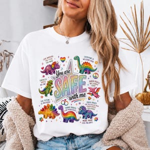 Può includere: Una t-shirt bianca con una grande stella centrale con il testo "You are SAFE with me" in colori arcobaleno scintillanti. Intorno alla stella ci sono dinosauri colorati, cuori e il testo "Love whoever the f*ck you want."