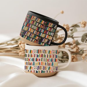 May include: Two ceramic mugs stacked vertically. The top mug is black with a colorful bookshelf pattern featuring rows of books and floral accents. The bottom mug is white with an identical bookshelf pattern. Both have a natural tan base.