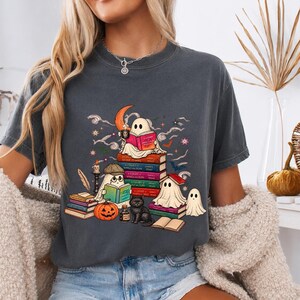 May include: A charcoal gray t-shirt featuring a graphic of ghosts reading books, a black cat, a pumpkin, and a crescent moon. The illustration includes stacks of colorful books with visible text, a candle, and stars, creating a detailed scene.