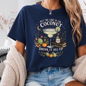 Könnte beinhalten: Ein marineblaues T-Shirt mit einem Grafikdruck, der ein Margaritaglas, eine Flasche mit der Aufschrift Midnight Elixir, Bücher und Früchte zeigt. Der Text lautet: Put the lime in the coconut, Midnight Margaritas und Drink it all up.