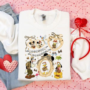 Pode incluir: Camisola branca com personagens de desenhos animados Mickey e Minnie Mouse, Pato Donald, Pateta e Pluto em trajes de safari. O design inclui o texto "Animal Kingdom" e molduras decorativas com estampas de leopardo.