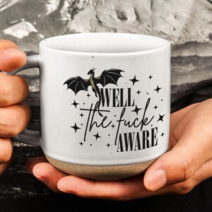 May include: A white ceramic mug with a speckled finish and a tan base. The front features a black graphic of a dragon above the text "WELL the fuck AWARE" surrounded by small black star icons. The mug has a curved handle on the left side.