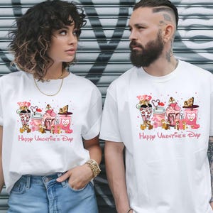 Puede incluir: Camisetas blancas con un gráfico colorido que presenta personajes de dibujos animados, postres y el texto "Happy Valentine's Day". El diseño incluye elementos rosas, marrones y rojos. Las camisetas las lleva una pareja.