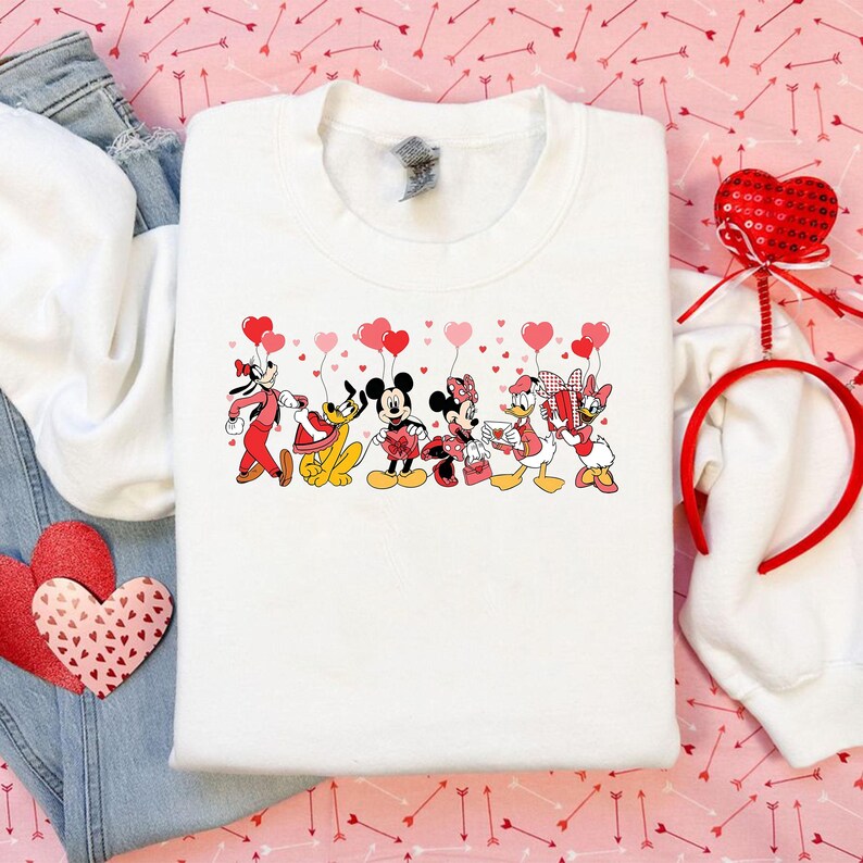 Pu&ograve; includere: Felpa bianca con una grafica di personaggi Disney, tra cui Topolino e Minnie, che tengono palloncini a forma di cuore. I personaggi sono vestiti con abiti di San Valentino. Sono visibili anche una fascia e una decorazione a forma di cuore rossi.