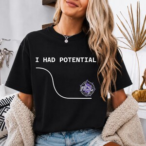 Può includere: Una t-shirt nera a maniche corte con il testo bianco I HAD POTENTIAL sopra un grafico a linee bianco che curva verso il basso verso una forma geometrica viola e bianca che ricorda un dodecaedro con una piccola figura all'interno.