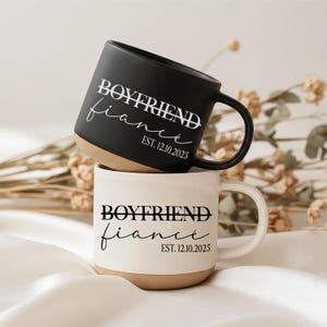 Puede incluir: Una taza de cerámica negra apilada sobre una taza de cerámica blanca moteada. Ambas tazas presentan el texto "BOYFRIEND" con una línea tachada, seguido de la palabra "fiancé" en cursiva y "EST. 12.10.2025" en una fuente sans-serif.