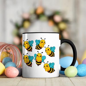 Könnte beinhalten: Weißer Keramikbecher mit schwarzem Henkel und Rand, verziert mit Cartoon-Bienen. Die Bienen sind gelb und schwarz mit blauen Flügeln. Der Becher steht auf einer Holzoberfläche mit bunten Ostereiern.