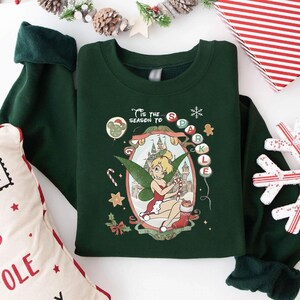 Könnte beinhalten: Dunkelgrünes Sweatshirt mit einem weihnachtlichen Tinkerbell-Motiv. Das Design umfasst ein Schloss, eine Zuckerstange und den Schriftzug "'Tis the season to sparkle". Das Sweatshirt hat dunkelgrüne Bündchen.