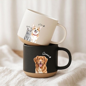 Könnte beinhalten: Ein cremefarbener Keramikbecher mit einer Illustration einer grauen Katze und eines Corgis mit den Namen Bella und Mila, gestapelt auf einem mattschwarzen Keramikbecher mit einem Golden Retriever und dem Namen Goose.
