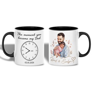 Op de afbeelding: Witte keramische mok met een zwarte binnenkant en handvat. Eén kant heeft een klokontwerp met de tekst "The moment you became my Dad" en de datum 02.01.2026. De andere kant toont een aquarel illustratie van een vader die een baby vasthoudt.