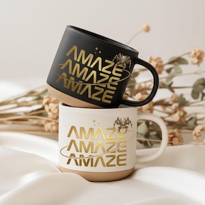 May include: Two ceramic mugs, one black and one white, stacked on top of each other. The black mug has gold lettering that says "AMAZE" and a gold spider graphic. The white mug has the same design. Both mugs have a tan base.
