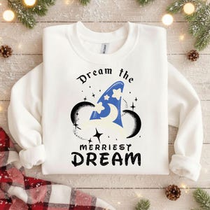 Könnte beinhalten: Weißes Sweatshirt mit einem Aufdruck, der einen blauen Zaubererhut mit Sternen und die Worte "Dream the Merriest Dream" in Schwarz zeigt. Das Design umfasst schwarze Mickey Mouse Ohren und Sternakzente. Ein gemütliches Oberteil mit langen Ärmeln.