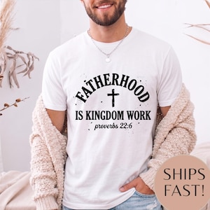 Könnte beinhalten: Weißes T-Shirt mit dem schwarzen Text "FATHERHOOD IS KINGDOM WORK" und einem Kreuzsymbol. Das Shirt enthält auch den Bibelvers "Sprüche 22,6". Das Shirt wird von einer Person getragen.