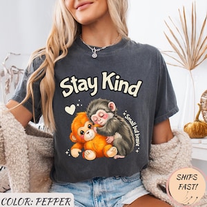 Op de afbeelding: Donkergrijs t-shirt met de witte tekst "Stay Kind", met een afbeelding van twee cartoonapen die elkaar omhelzen. Eén aap is oranje, de andere is grijs. De tekst "Small but brave" staat ook op het shirt.
