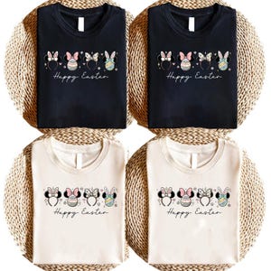Può includere: Quattro magliette, due nere e due color crema, ciascuna con una grafica a tema pasquale. Il design include orecchie di Minnie Mouse, uova di Pasqua e la scritta "Happy Easter" in un font script. Le magliette sono esposte su una superficie intrecciata.