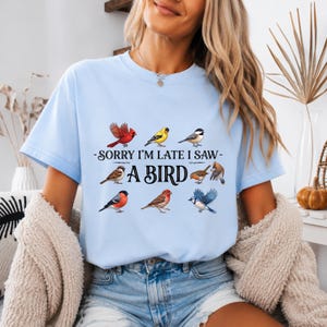 Könnte beinhalten: Ein hellblaues T-Shirt mit einem Grafikdruck verschiedener bunter Vögel, die um den schwarzen Text Sorry I'm Late I Saw A Bird angeordnet sind. Das Design enthält Illustrationen eines Kardinals, Stieglitz, Meise, Sperlings, Rotkehlchens und Blauhähers.