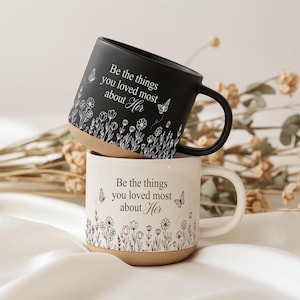 Könnte beinhalten: Zwei Keramikbecher, einer in Mattschwarz und einer in Off-White, jeweils mit einem beigefarbenen Boden. Beide Becher zeigen den Text 'Be the things you loved most about Her' in Serifenschrift, umgeben von Strichzeichnungen von Wildblumen und Schmetterlingen.