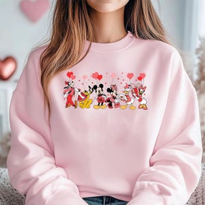 Pu&ograve; includere: Felpa rosa chiaro con una grafica di personaggi Disney, tra cui Topolino e Minnie, che tengono in mano palloncini a forma di cuore. I personaggi indossano abiti di San Valentino. La felpa &egrave; di colore rosa chiaro.