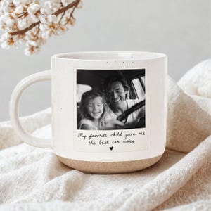 May include: A cream-coloured ceramic mug with a speckled finish and a rounded handle. The front features a square black and white photograph of a child and an adult in a car, with the text "My favorite child gave me the best car rides" below.