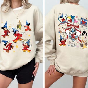Könnte beinhalten: Beiges Sweatshirt mit Mickey Mouse im Zauberer-Outfit. Auf der Vorderseite ist Mickey in verschiedenen Posen mit Sternen zu sehen. Auf der Rückseite ist Mickey mit einem herzförmigen Design, Luftballons und Schlossillustrationen abgebildet.