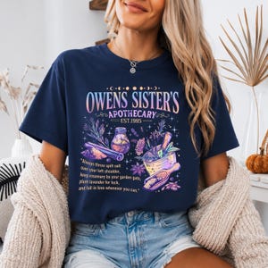 Könnte beinhalten: Ein marineblaues T-Shirt mit einem Aufdruck, der den Text "OWENS SISTER'S APOTHECARY EST. 1995" sowie ein Zitat über Salz, Rosmarin und Lavendel zeigt. Das Design enthält Illustrationen von Mörser und Stößel, einem Glas und Zimtstangen.