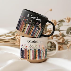 Könnte beinhalten: Zwei Keramikbecher mit Blumen- und Buchdesign. Ein Becher ist schwarz, der andere cremefarben. Beide Becher tragen den Namen "Madeline" in Schreibschrift. Die Becher zeigen eine Reihe bunter Bücher mit Blumen.
