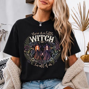 Pode incluir: Uma t-shirt preta de manga curta com uma estampa gráfica de duas figuras com chapéus de bruxa e vestidos escuros segurando guarda-chuvas, cercadas por uma coroa de flores. O texto diz, There is a little WITCH In all of us, em uma fonte decorativa.