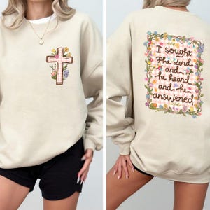 Può includere: Felpa beige con un disegno a croce sul davanti e testo sul retro. La croce è decorata con un motivo a quadri rosa e dettagli floreali. Il retro della felpa presenta la scritta "I sought The Lord and he heard and he answered" circondata da fiori.
