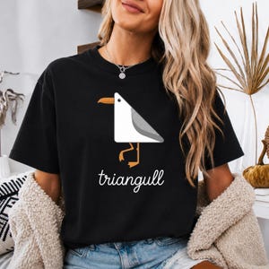 Könnte beinhalten: Ein schwarzes Kurzarm-T-Shirt mit einer Grafik einer weißen und grauen Möwe in Dreiecksform, die einen orangefarbenen Schnabel und Füße hat. Unter dem Vogel ist das Wort triangull in weißer Schreibschrift auf der Vorderseite aufgedruckt.