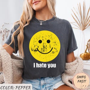 Könnte beinhalten: Pfefferfarbenes T-Shirt mit einem verblichenen gelben Smiley-Gesicht und dem weißen Text "I hate you". Das Shirt hat kurze Ärmel und einen Rundhalsausschnitt.