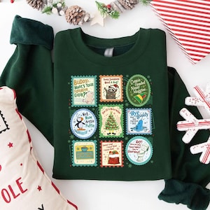 Könnte beinhalten: Ein dunkelgrünes Sweatshirt mit einer Sammlung farbenfroher, stempelförmiger Grafiken. Die Grafiken zeigen Bilder eines Eisbären, eines Waschbären, eines Weihnachtsbaums und eines Narwals sowie Text wie "Buddy, What's Your Favorite Color?" und "Cotton Headed Ninny Muggins."