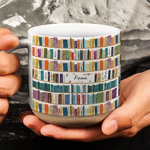 Könnte beinhalten: Ein weißer Keramikbecher mit gesprenkeltem beigefarbenem Boden und einem wiederkehrenden Muster aus bunten Büchern in Regalen. Das Design zeigt verschiedene Buchrücken in Blau-, Grün-, Orange- und Violetttönen sowie den Namen Anna in schwarzer Schrift.