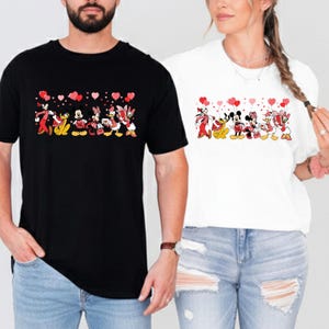 Pu&ograve; includere: T-shirt bianche e nere con un design a cartoni animati di personaggi Disney che tengono in mano palloncini a forma di cuore. I personaggi includono Topolino, Minnie, Paperino, Paperina e Pippo. Le magliette sono indossate da due persone.