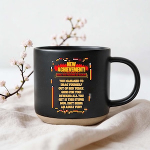 Könnte beinhalten: Schwarze Kaffeetasse mit beigefarbenem Boden, mit dem Text "NEW ACHIEVEMENT!" und einer humorvollen Botschaft über das Erwachsenenleben. Das Design der Tasse umfasst pixelige Grafiken in Rot, Gelb und Weiß.