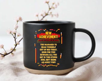 May include: Black coffee mug with a tan base, featuring the text "NEW ACHIEVEMENT!" and a humorous message about adulting. The mug's design includes pixelated graphics in red, yellow, and white.