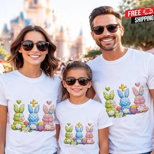 Könnte beinhalten: Drei Personen tragen weiße T-Shirts mit einem farbenfrohen Osterhasen-Design. Das Design umfasst grüne, blaue und rosa Hasen, ein Kreuz, Herzen und Ostereier. Die Personen lächeln und tragen Sonnenbrillen.