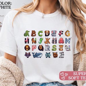 Könnte beinhalten: Weißes T-Shirt mit farbenfrohem Tieralphabet. Das Shirt hat den Text "COLOR: WHITE" oben links und "SUPER SOFT TEES!" unten rechts. Die Buchstaben sind mit Tierillustrationen gepaart.