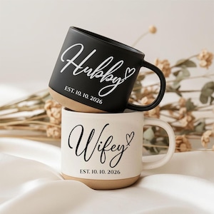 Op de afbeelding: Een zwarte keramische mok met de witte tekst Hubby en EST. 10. 10. 2026 gestapeld op een witte keramische mok met de zwarte tekst Wifey en EST. 10. 10. 2026. Beide mokken hebben een hartpictogram en een natuurlijke beige basis met spikkels.