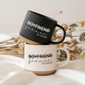 Könnte beinhalten: Zwei Keramikbecher, einer schwarz und einer weiß, übereinander gestapelt. Der schwarze Becher hat die Worte "BOYFRIEND fiancé" und "EST. 12.10.2025" in Weiß aufgedruckt. Der weiße Becher hat denselben Text in Schwarz. Beide Becher haben einen beigefarbenen Boden.